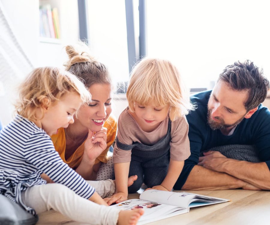 Front view of young family with two small children indoors in bedroom reading a book.